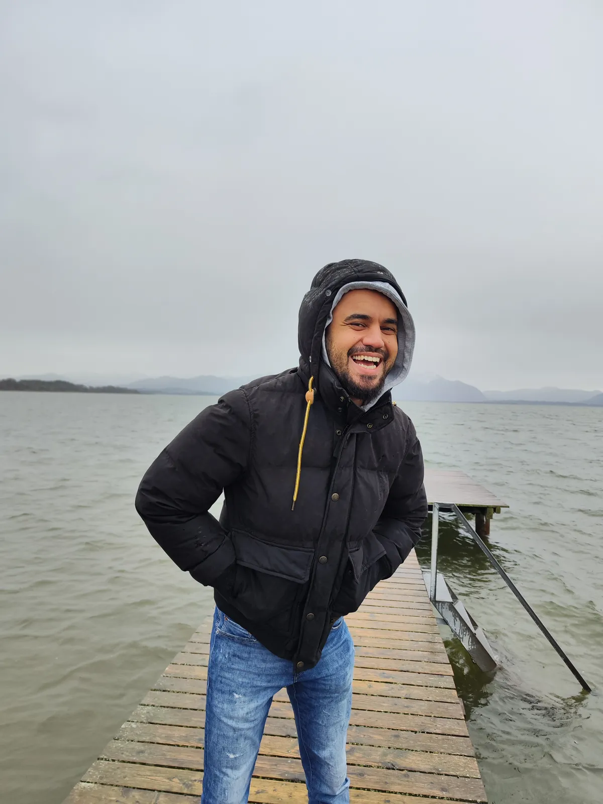 Ashkan Vahdani lachend auf einem Holzsteg am Chiemsee, im Hintergrund die Berge